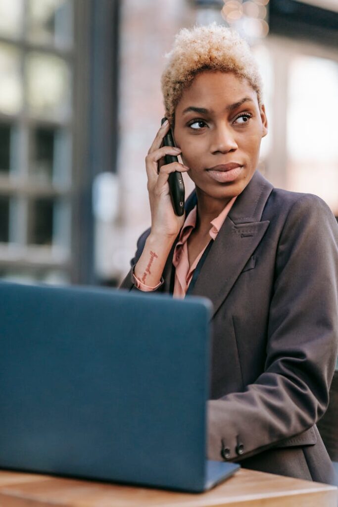 Black woman talking on smartphone and browsing laptop for work. She is a freelance project manager for ResultsResourcing.