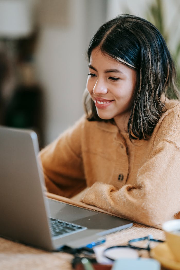 Young cheerful Latin American female remote worker browsing internet on netbook at table at home