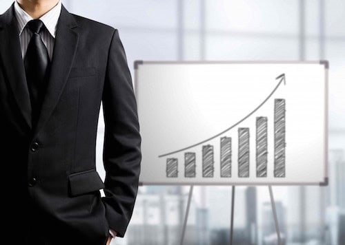 A man is standing in front of a bar graph with an upward-facing​ arrow suggesting business growth and success in scaling his business. 